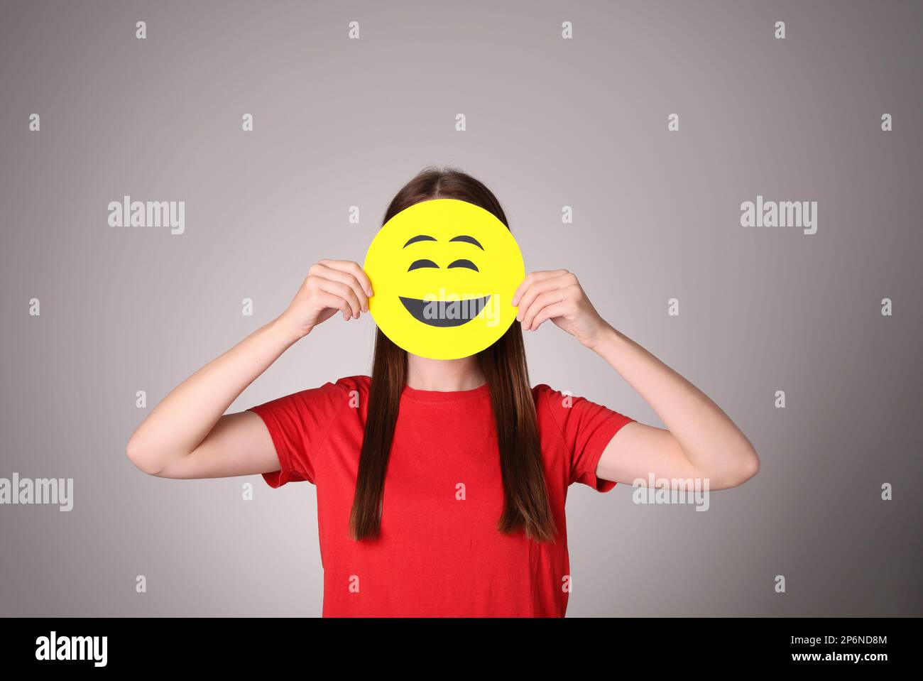 Woman covering face with laughing emoji on grey background Stock Photo