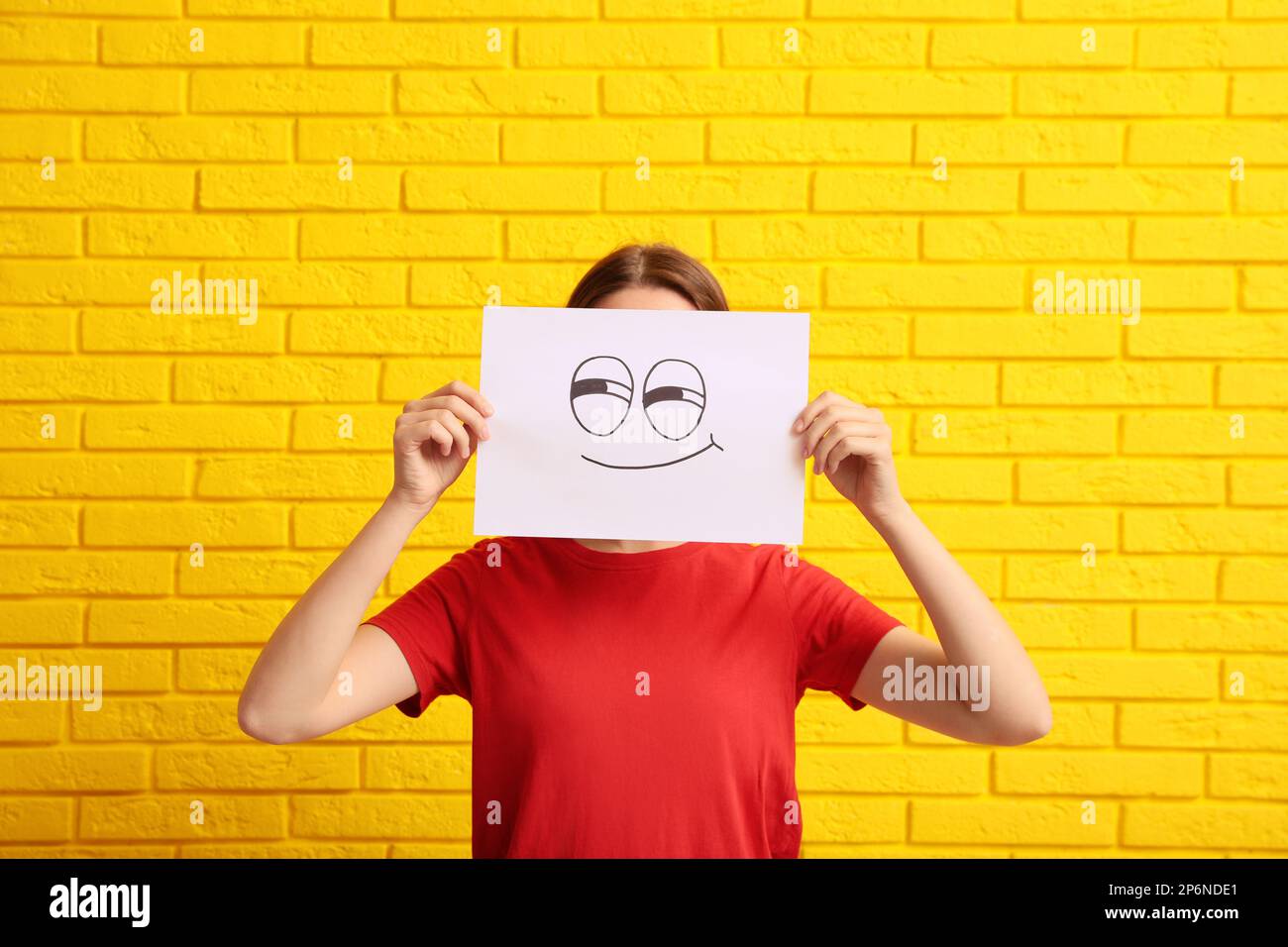Woman hiding behind sheet of paper with smirking face near yellow brick wall Stock Photo