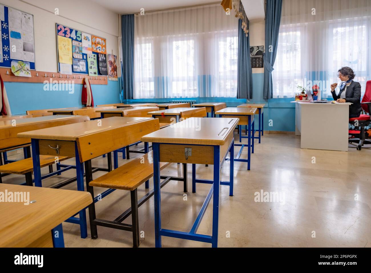 Empty class room with wood tables and female teacher sitting at her desk, reading notes in her notebook. Horizontal indoors shot with copy space. Stock Photo