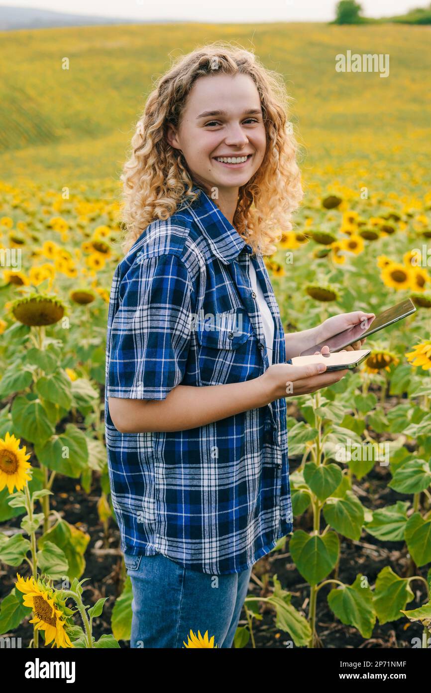 Smiling curly-haired farmer woman observing and recording information about quality of sunflower field, holding smart-phone and tablet Concept for Stock Photo