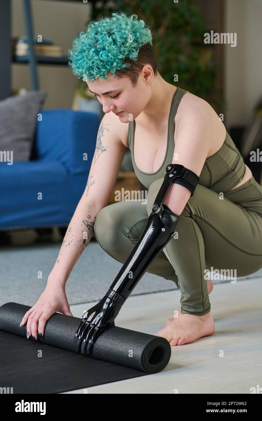 Vertical image of young woman with prosthetic arm preparing exercise mat for sport training at home Stock Photo