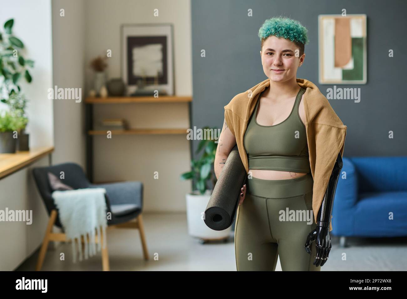 Portrait of young woman with prosthetic arm holding exercise mat for yoga and looking at camera while standing in the room Stock Photo