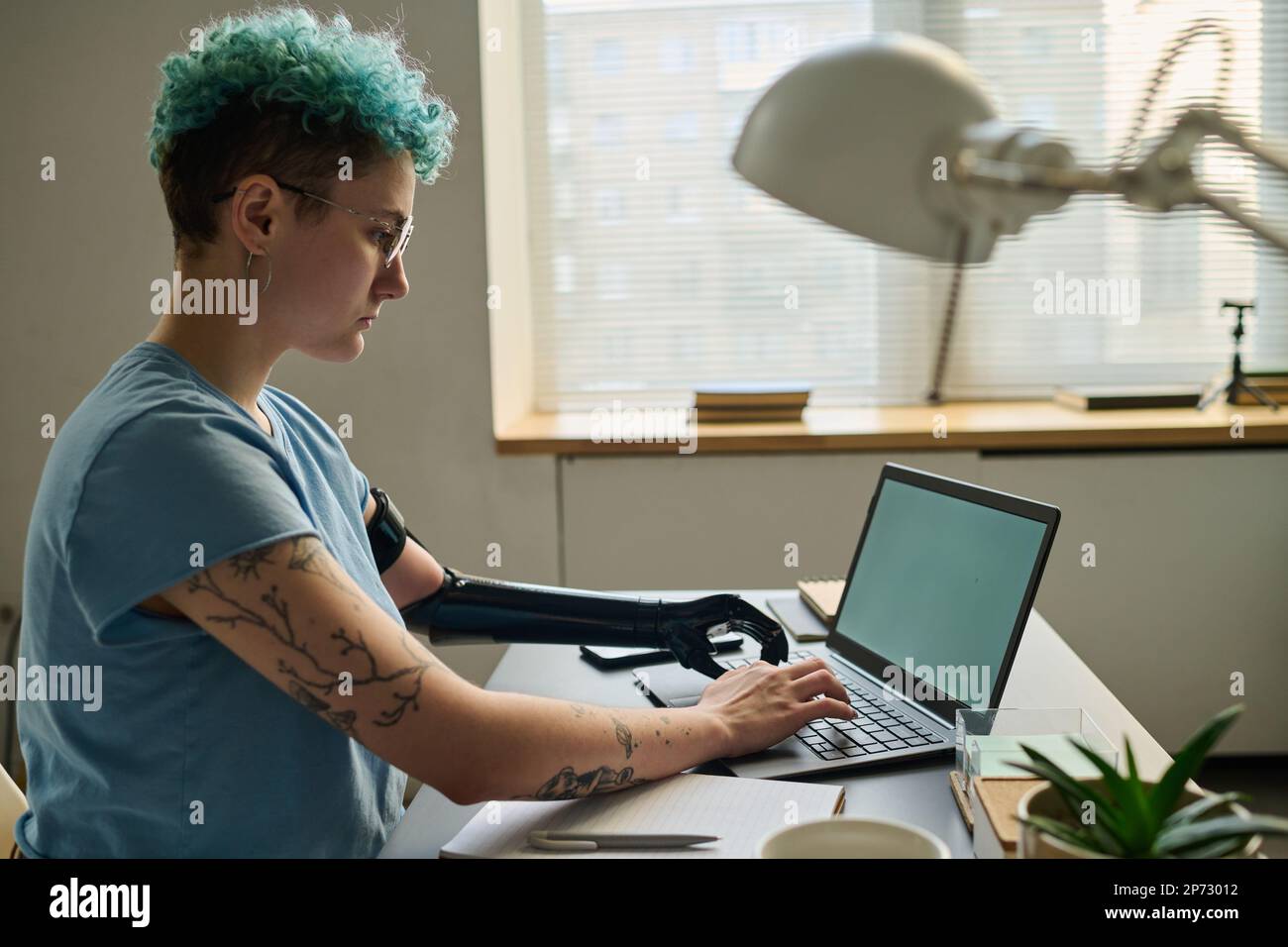 Young woman with prosthetic arm typing on laptop and working online at table in the room Stock Photo