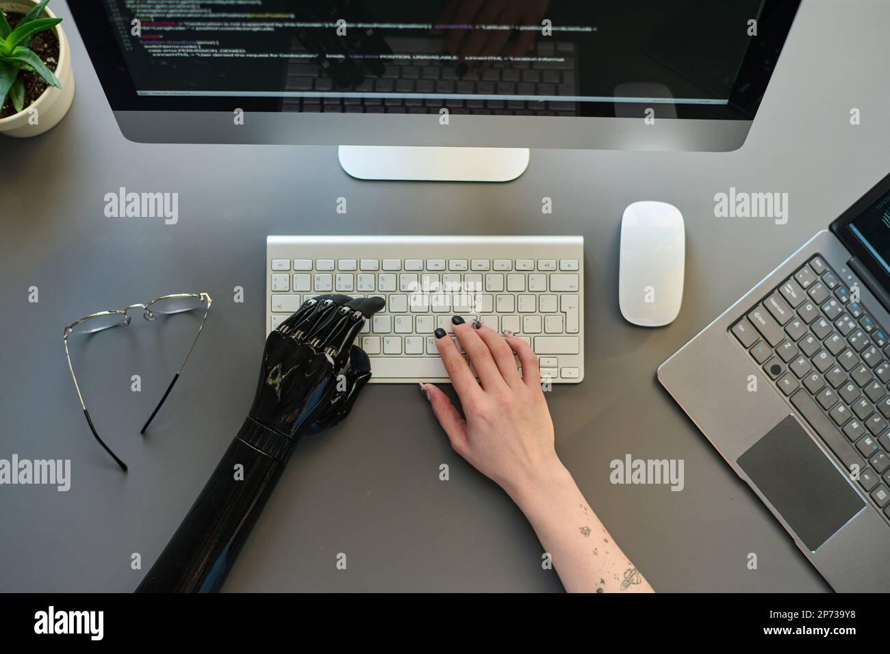 High angle view of young woman with prosthetic arm sitting in front of monitor, typing on keyboard and working with security codes Stock Photo