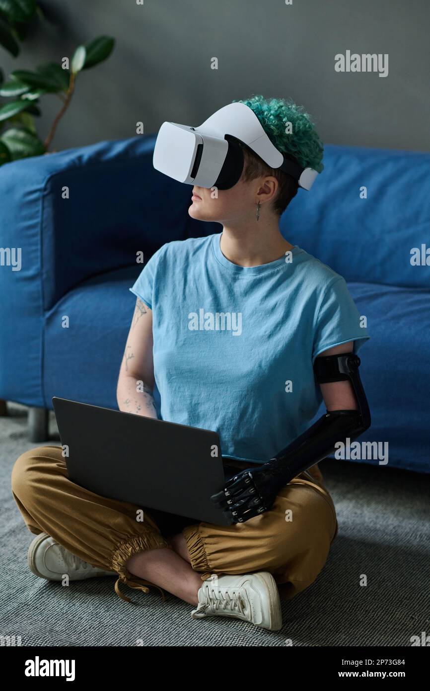 Woman with prosthetic arm in VR glasses playing in virtual reality game using laptop while sitting on the floor in the room Stock Photo