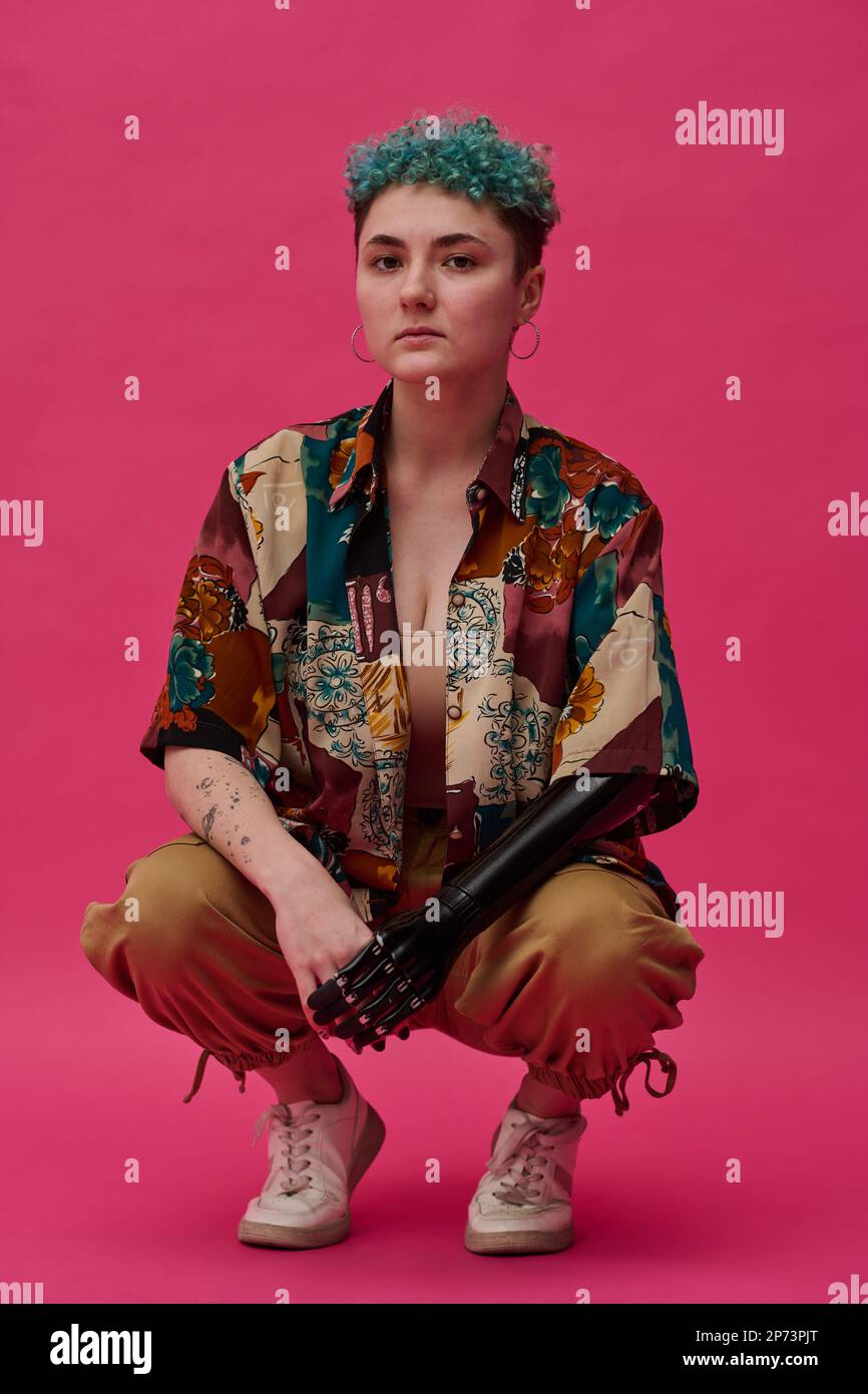 Portrait of young fashion girl with prosthetic arm loking at camera sitting against pink background Stock Photo