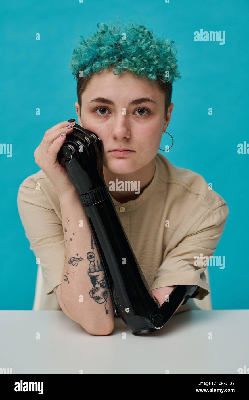Portrait of stylish teenager with prosthetic arm looking at camera sitting against blue background Stock Photo