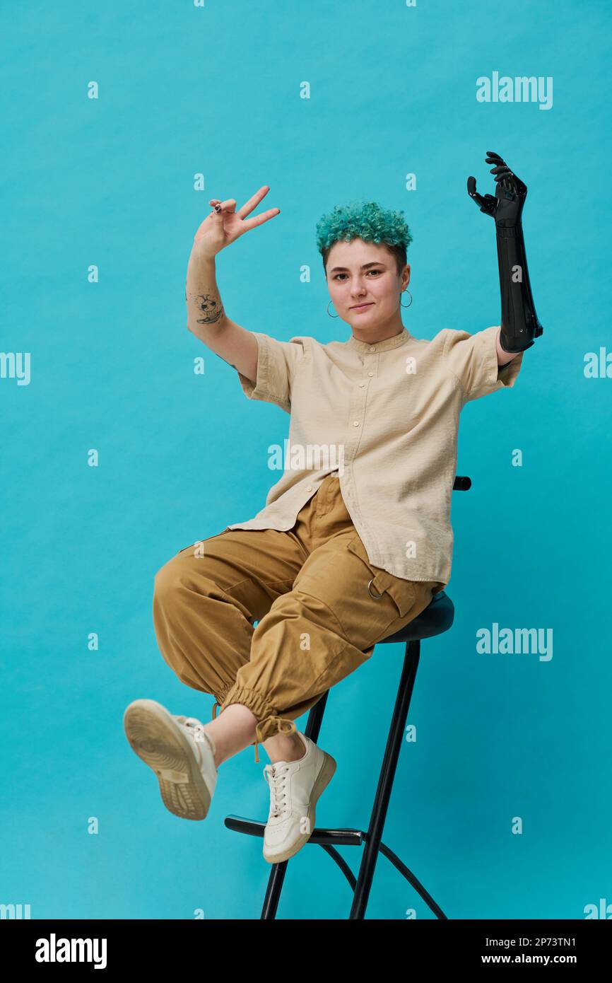Vertical image of teenage girl with prosthetic arm sitting on chair against blue background Stock Photo