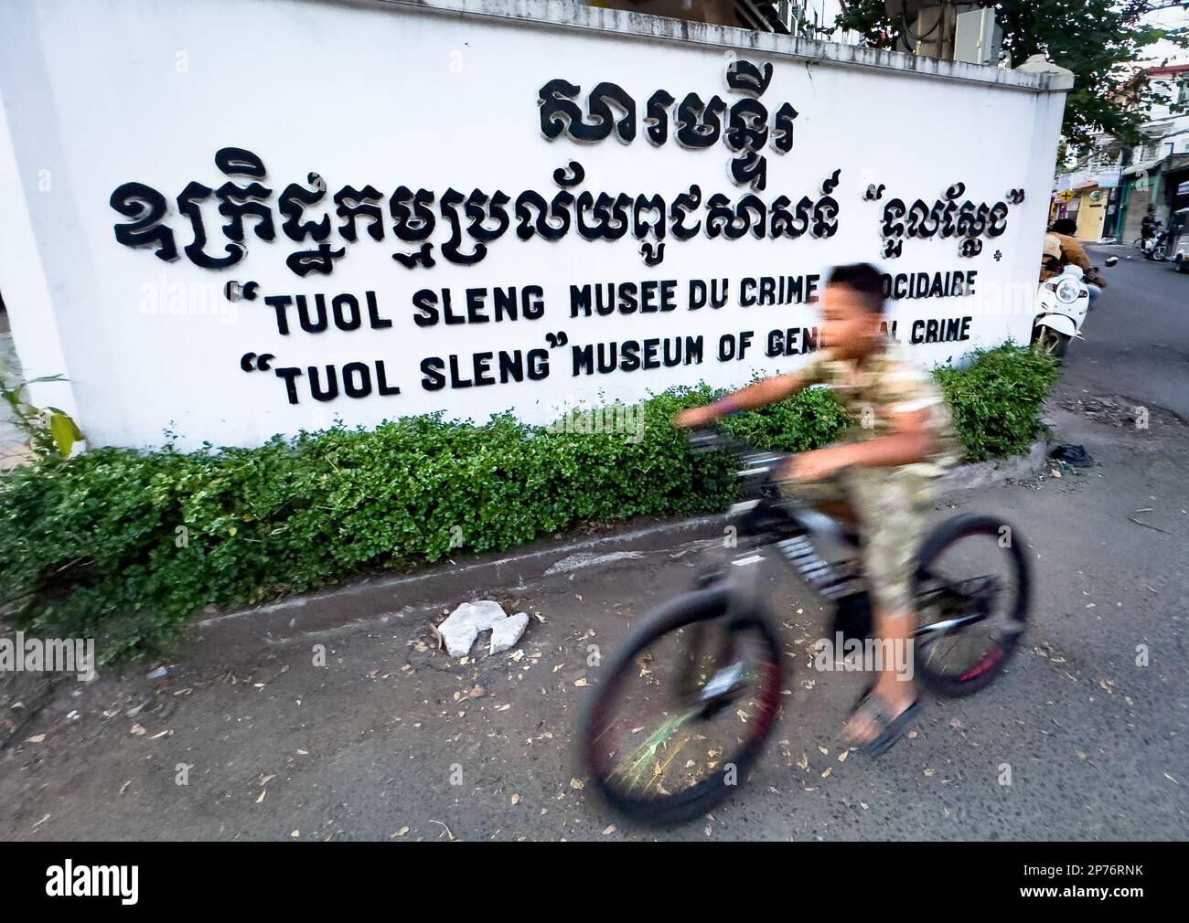 A young boy cycles past the entrance sign for the museum at the former Khmer Rouge torture death camp Tuol Sleng in Phnom Penh, Cambodia. Stock Photo