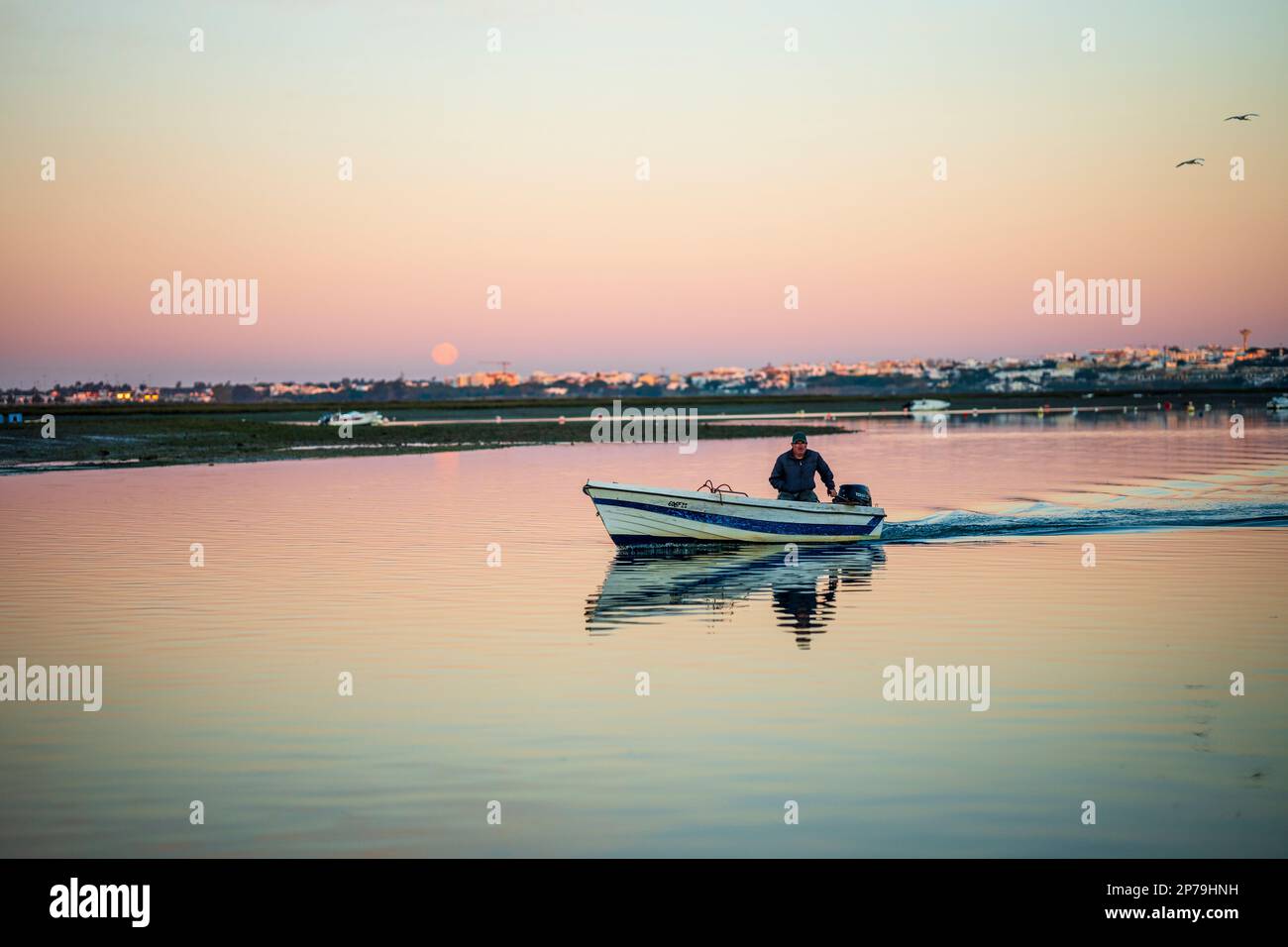 Faro, Portugal - February 5, 2023: A man traveling in his boat on Ria Formosa waters by sunrise and moonset Stock Photo