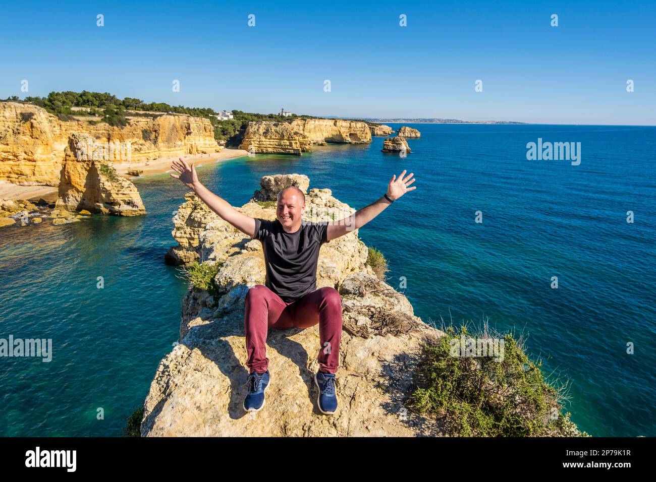 Happy man sitting with hands up on the top of cliffs at Marinha beach, Algarve, Portugal Stock Photo