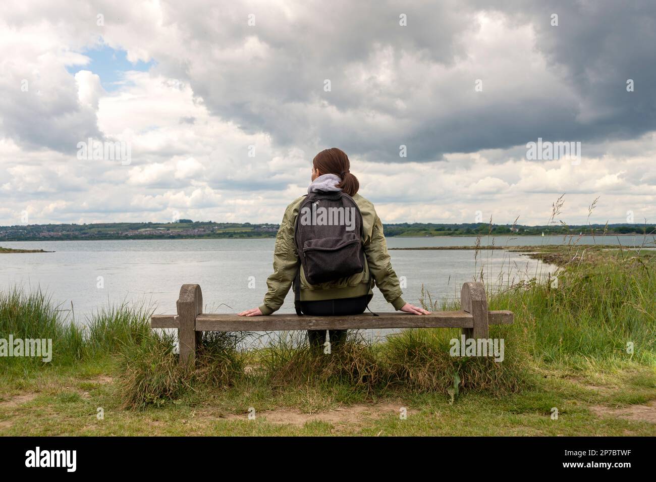 Woman sitting on a bench resting and enjoying the view across water. Thinking spot. Stock Photo