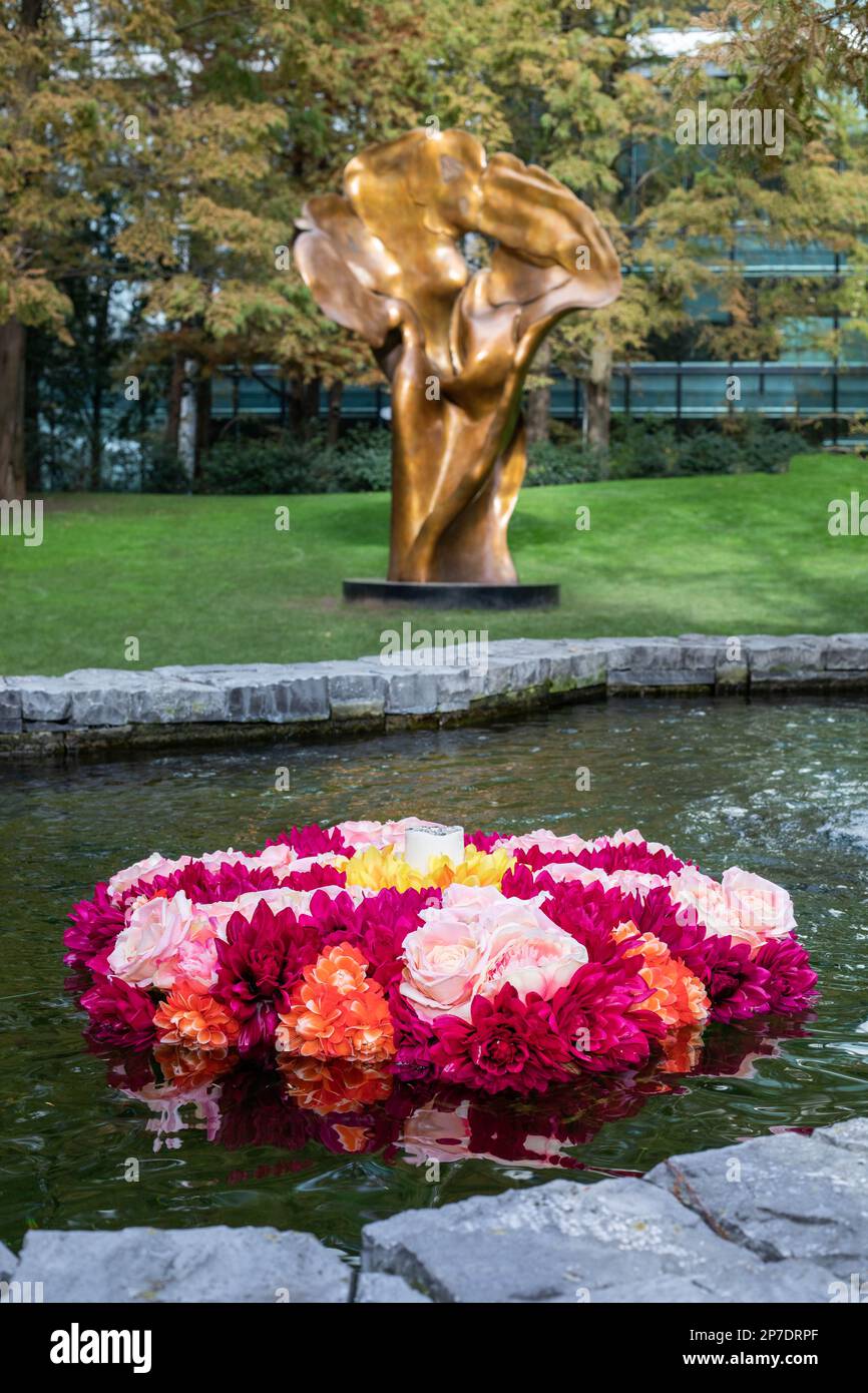 Colourful rangoli flowers float in a pool of water in front of the bronze artwork titled Fortuna by Helaine Blumenfeld in Jubilee Park, Canary Wharf Stock Photo