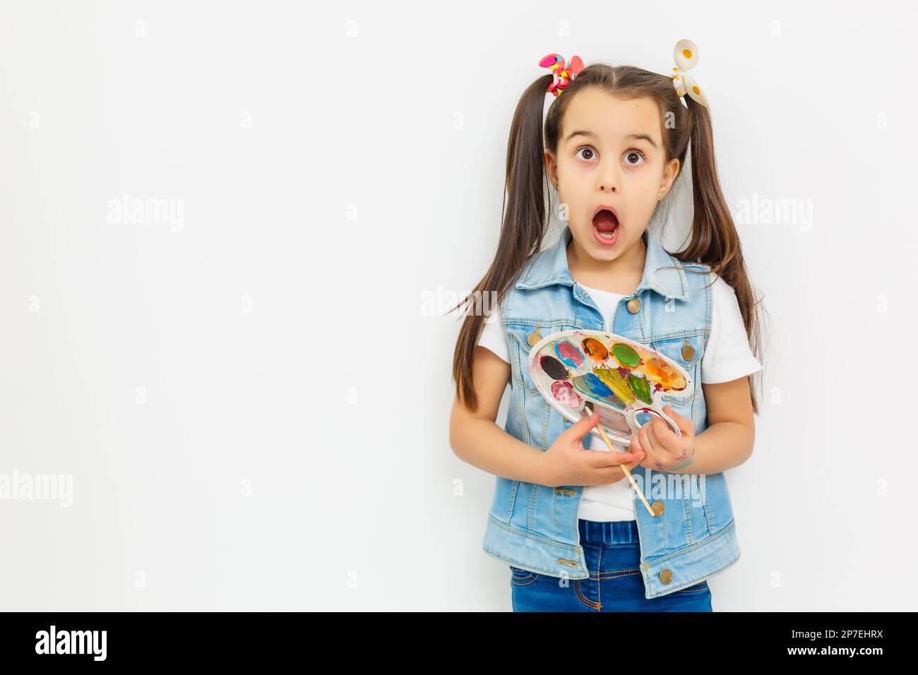 A joyful little girl with a paint palette and a brush on a white ...