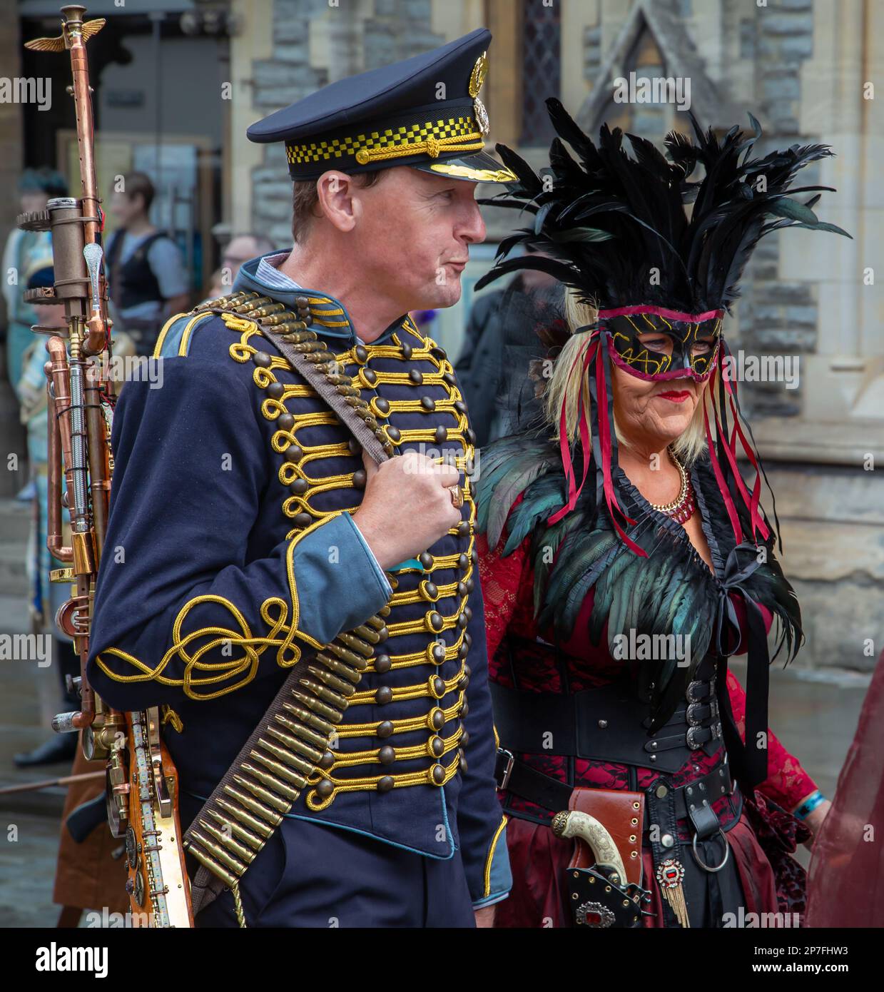 A male and female steampunk. The man is wearing a military style uniform and is carrying a weapon. The woman is wearing a feather headdress and a mask. Stock Photo