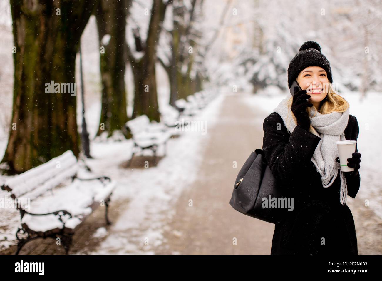 A young woman wearing warm winter clothes and a knit hat smiles happily as she stands in the snow and using mobile phone whil holding cofee cup Stock Photo