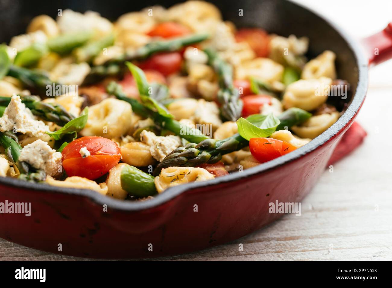 Cast iron pan with vegetable tortellini with asparagus, tomatoes, mushrooms and home made vegan feta cheese. Stock Photo