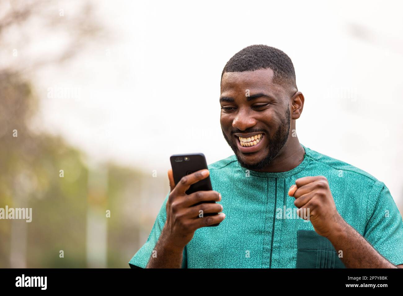 Excited Young African Man Holding smartphone outdoors, happy to win lottery. Stock Photo