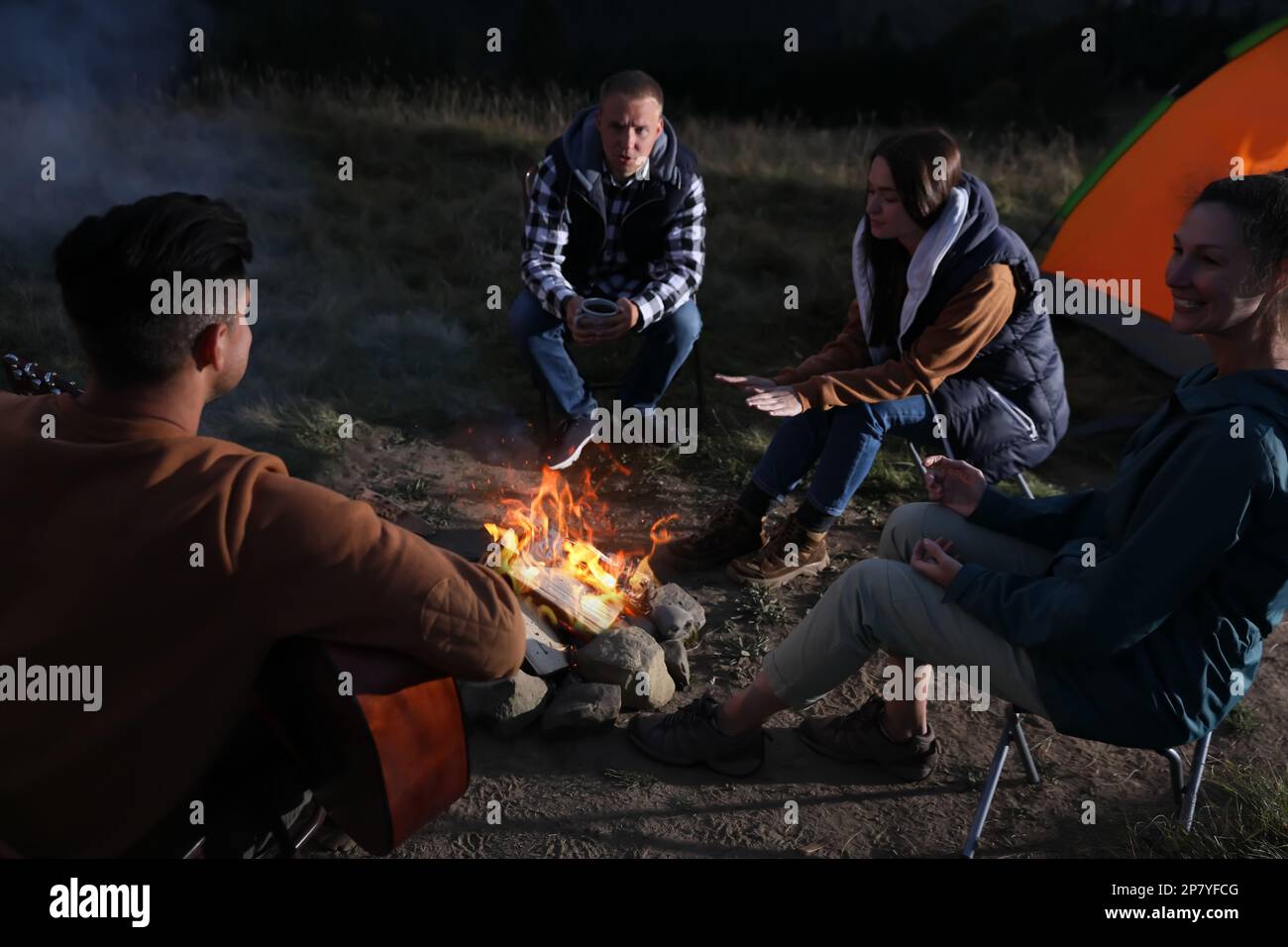 Group of friends gathering around bonfire at camping site in evening Stock Photo