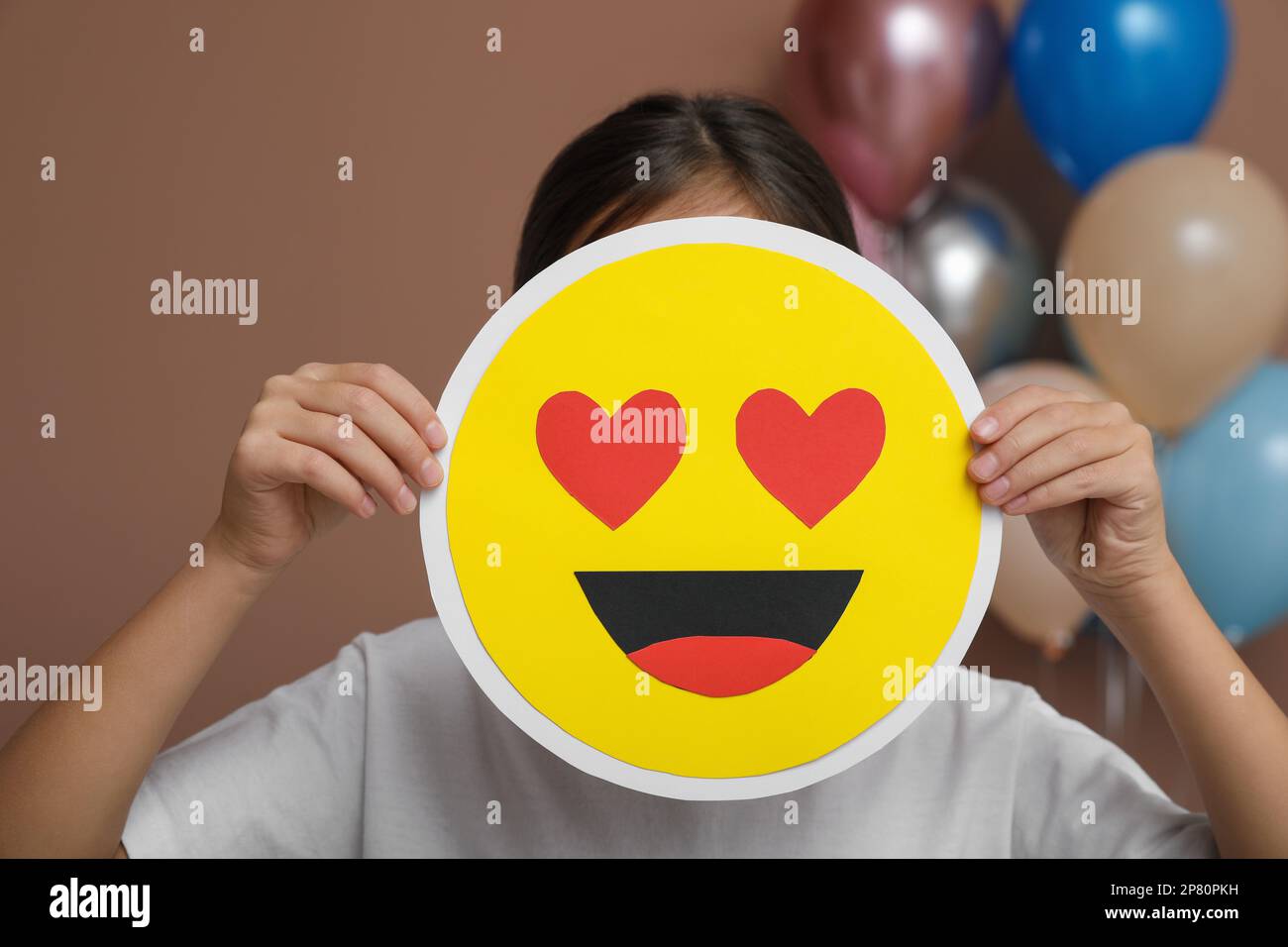 Little girl covering face with heart eyes emoji in decorated room Stock