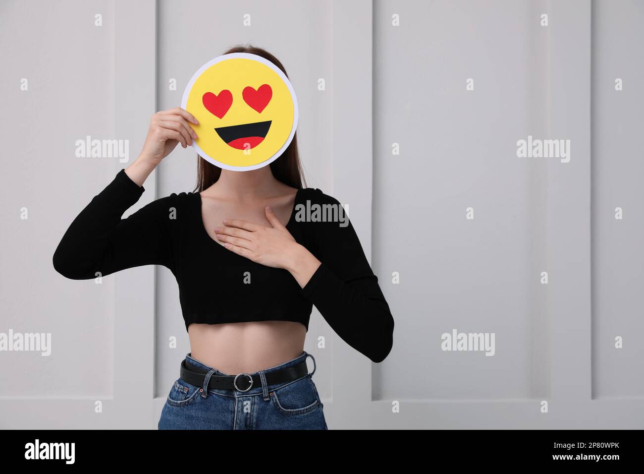 Woman covering face with heart eyes emoji near white wall, space for