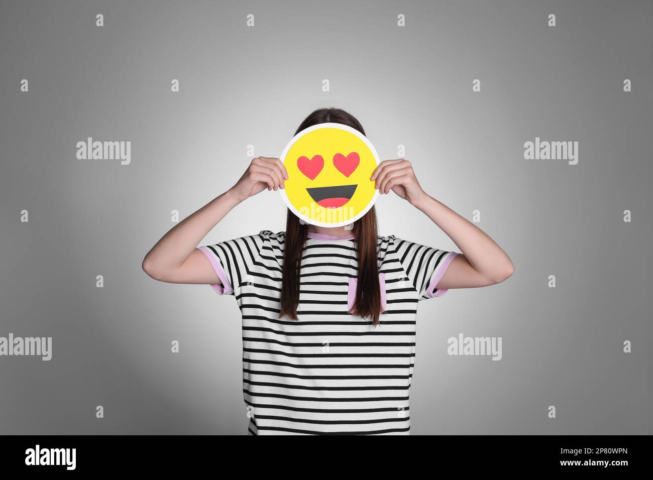 Woman covering face with heart eyes emoji on grey background Stock