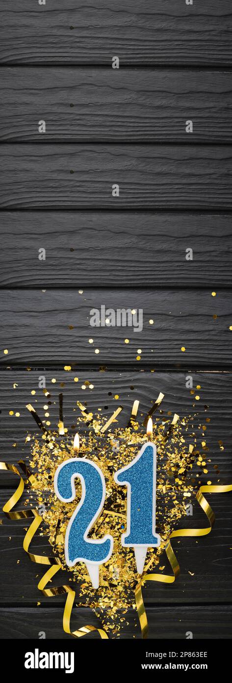 Number 21 blue celebration candle and gold confetti on dark wooden background. 21th birthday card. Anniversary and birthday concept. Vertical banner. Stock Photo