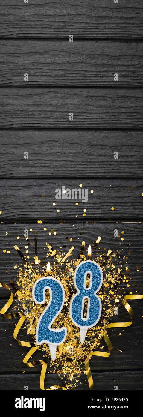 Number 28 blue celebration candle and gold confetti on dark wooden background. 28th birthday card. Anniversary and birthday concept. Vertical banner. Stock Photo