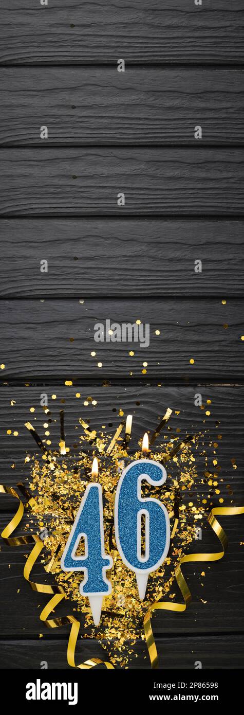 Number 46 blue celebration candle and gold confetti on dark wooden background. 46th birthday card. Anniversary and birthday concept. Vertical banner. Stock Photo