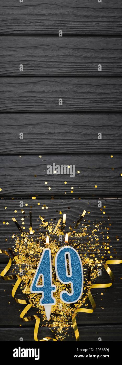 Number 49 blue celebration candle and gold confetti on dark wooden background. 49th birthday card. Anniversary and birthday concept. Vertical banner. Stock Photo