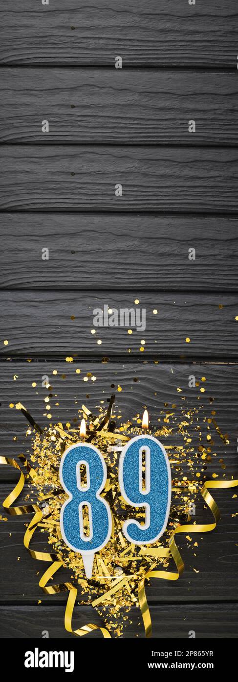 Number 89 blue celebration candle and gold confetti on dark wooden background. 89th birthday card. Anniversary and birthday concept. Vertical banner. Stock Photo