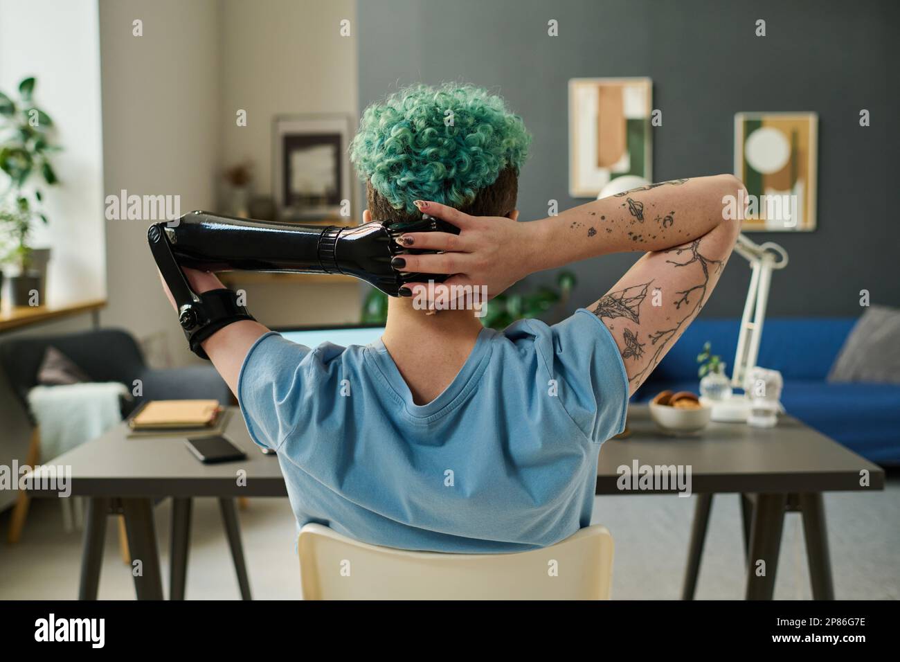 Rear view of young woman with prosthetic arm resting at table after online work in the room Stock Photo