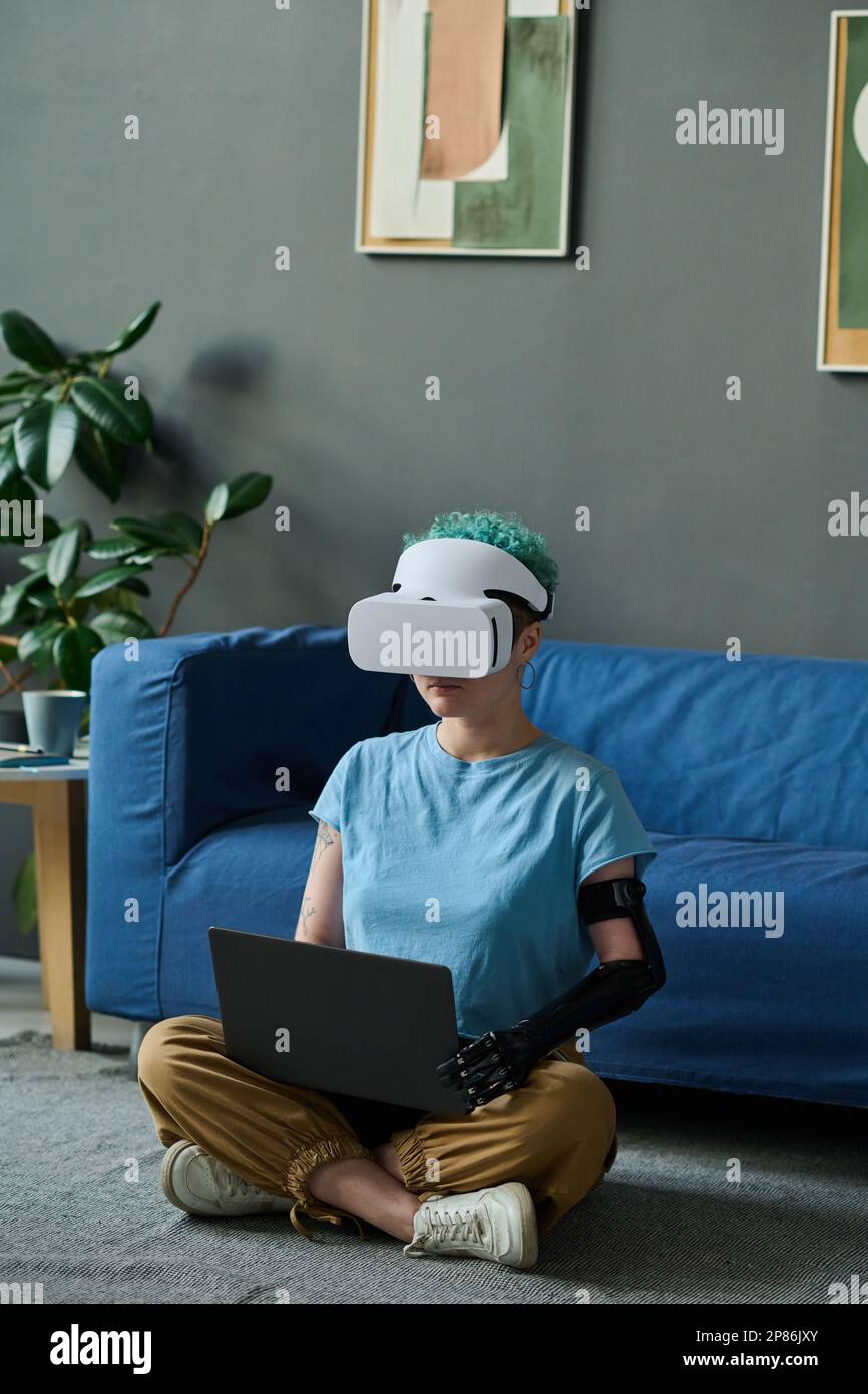 Young woman with prosthetic arm in VR glasses sitting on the floor in the room and using laptop Stock Photo