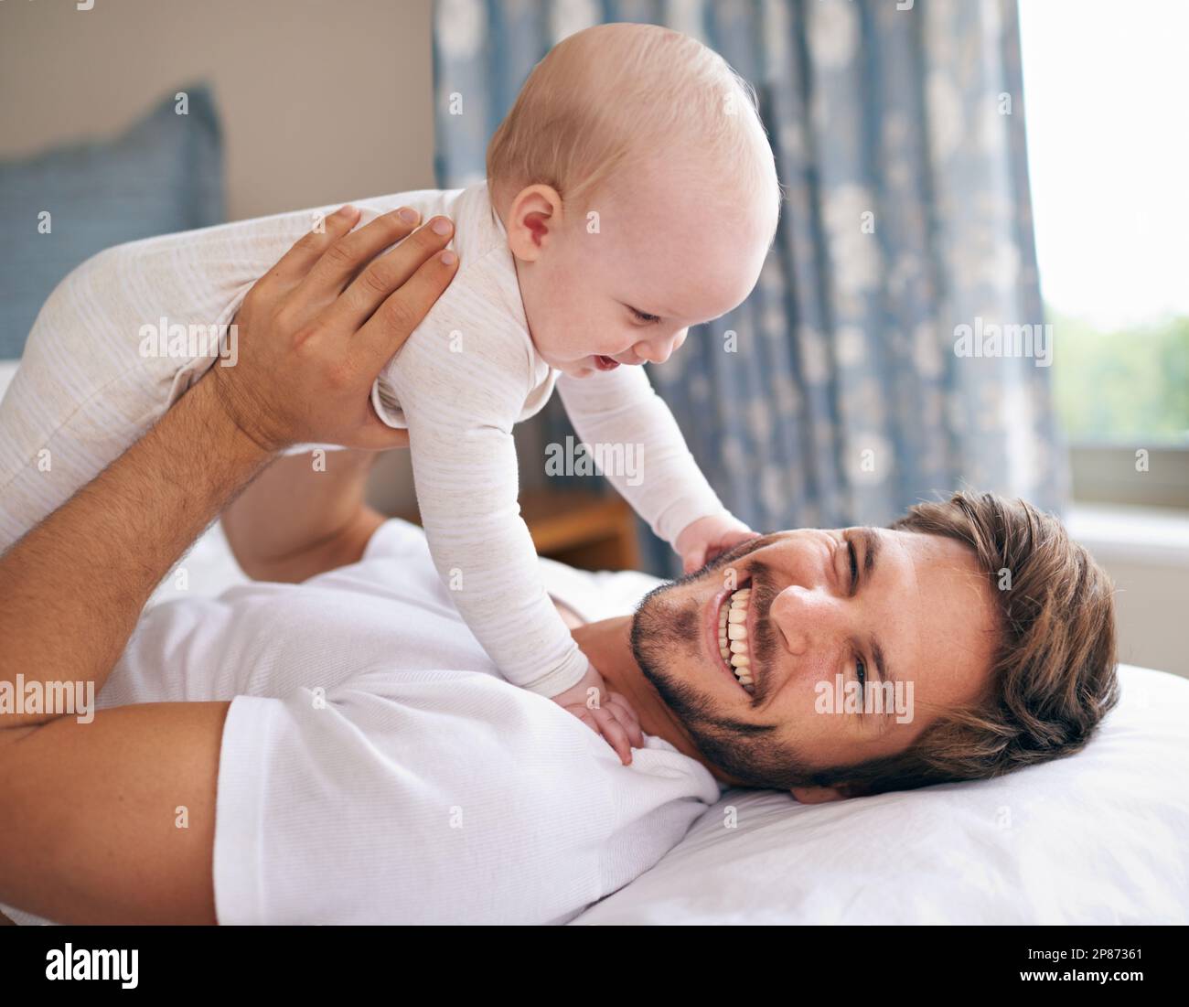 I love grabbing daddys beard. a father lying on a bed holding his baby girl up in the air. Stock Photo
