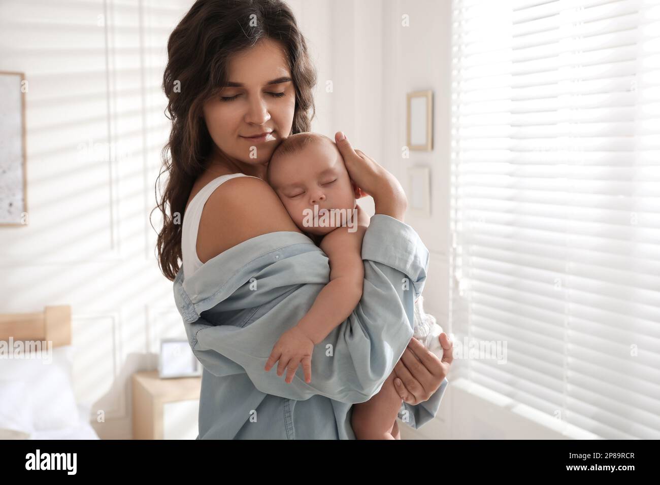 Happy young mother with her sleeping baby near window at home Stock