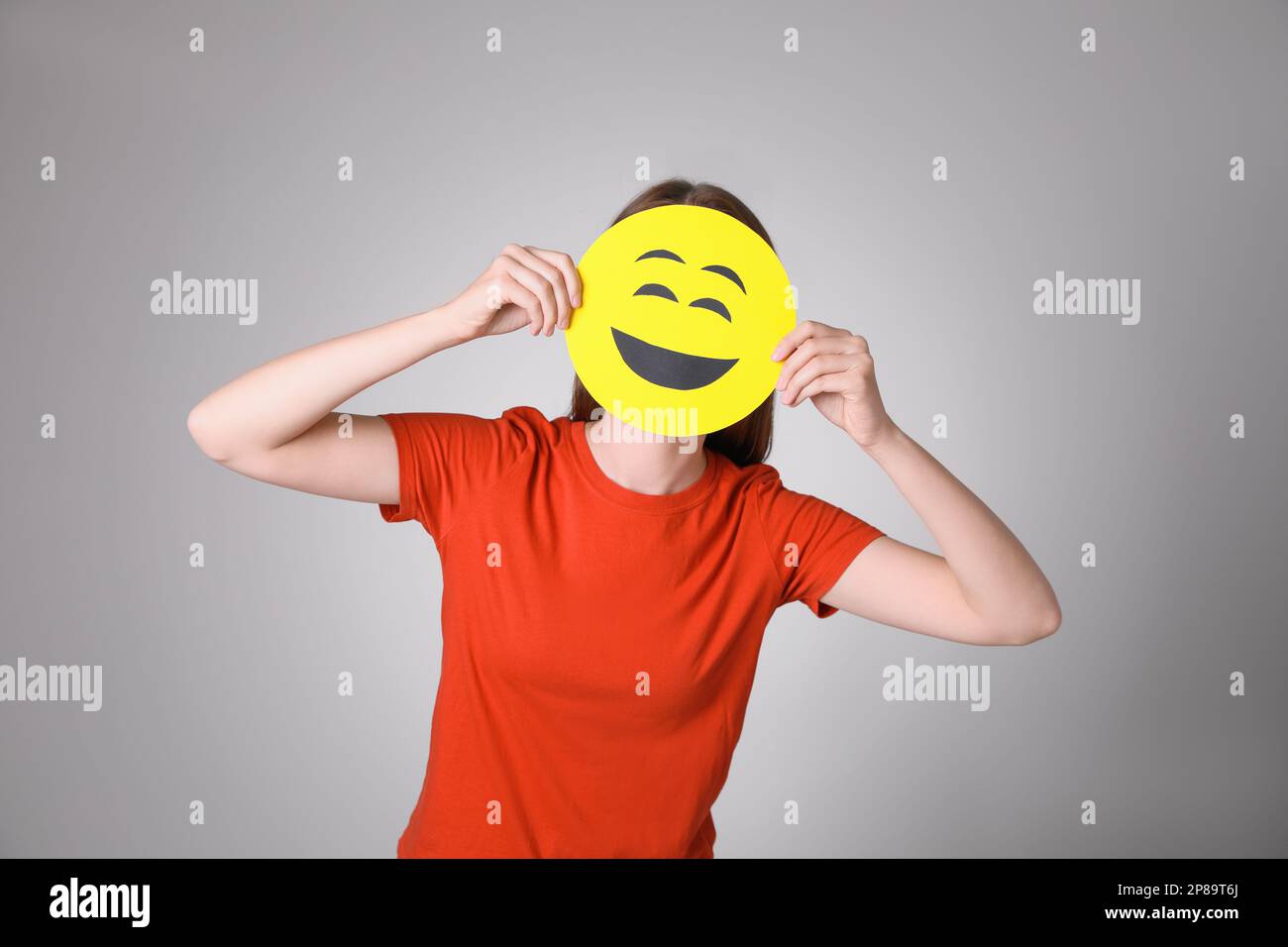 Woman covering face with laughing emoji on grey background Stock Photo