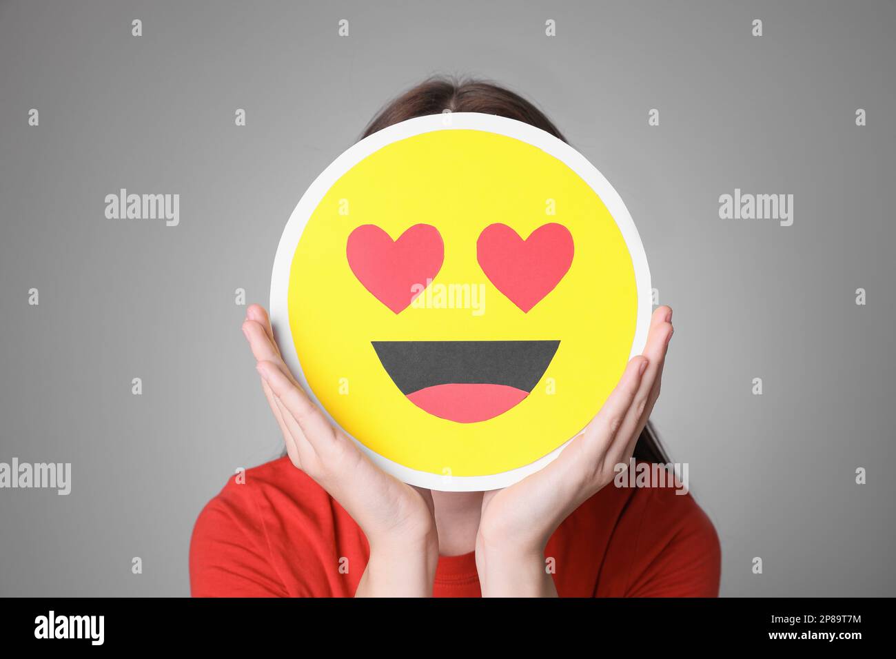 Woman covering face with heart eyes emoji on grey background Stock
