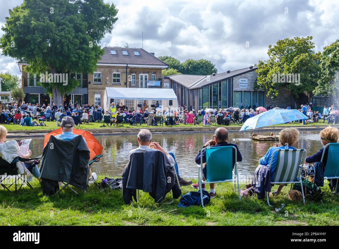 People sit on the ground & in chairs at Pinner Memorial park, watching