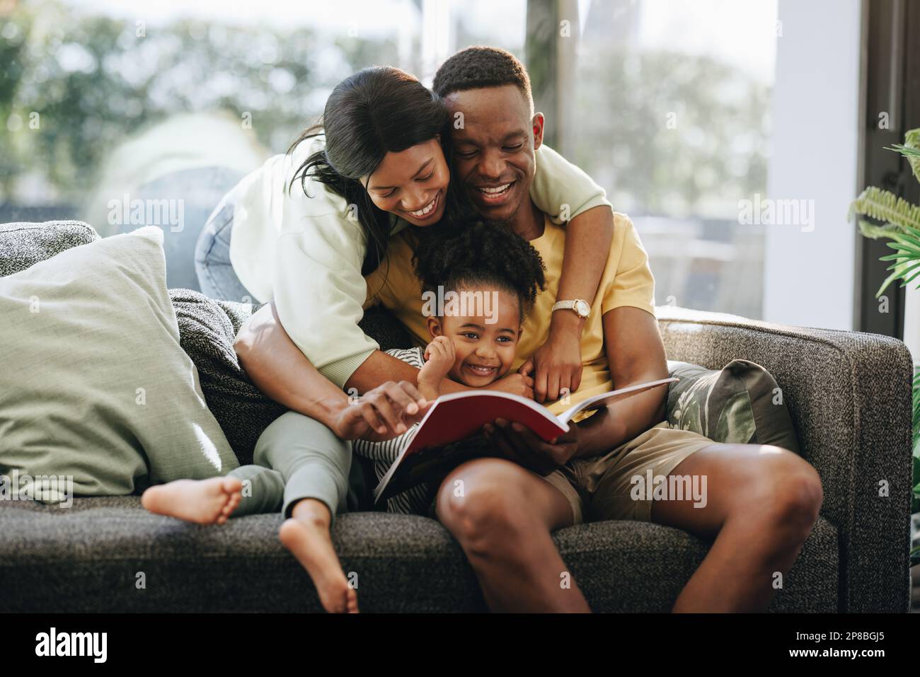 Parents reading a book with their daughter on a couch. Happy family spending time together at home. African couple raising a toddler in a loving envir Stock Photo