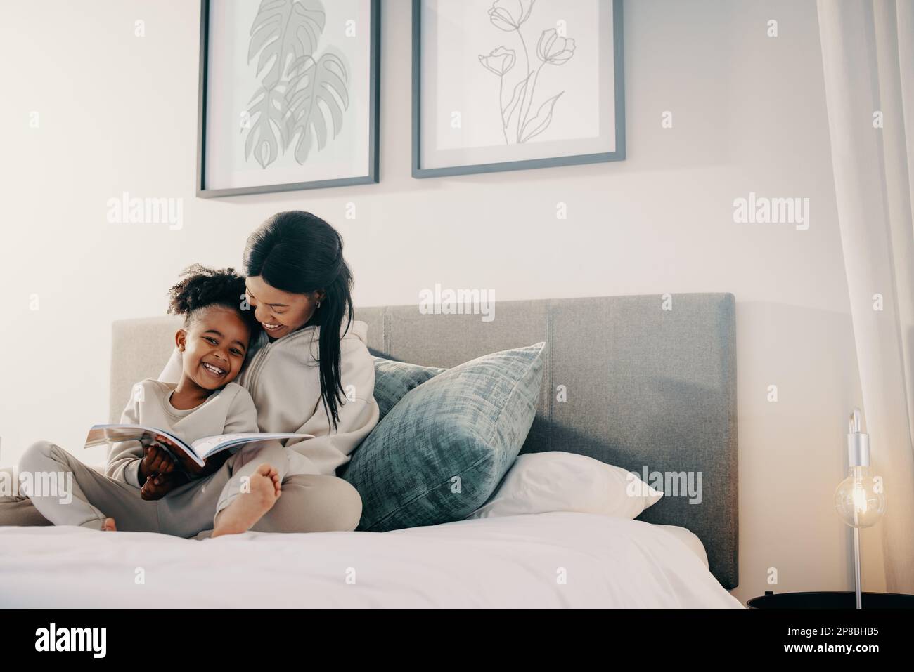 Mother and daughter spend time reading a book together, they’re smiling and sitting on a bed. Happy mom is helping her child with her reading skills. Stock Photo
