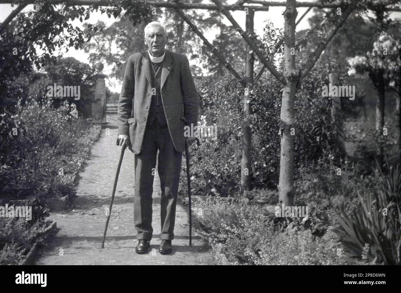 circa 1930s, historical, an elderly English vicar wearing a woollen three-piece suit and clerical collar, walking through a path in a garden with the aid of two walking sticks, one in each hand, England, UK. The wearing of a religious collar is a sign of the priest's availability, an identifying badge that can be recognised by people of all faiths and the permanent nature of Holy Orders. Stock Photo