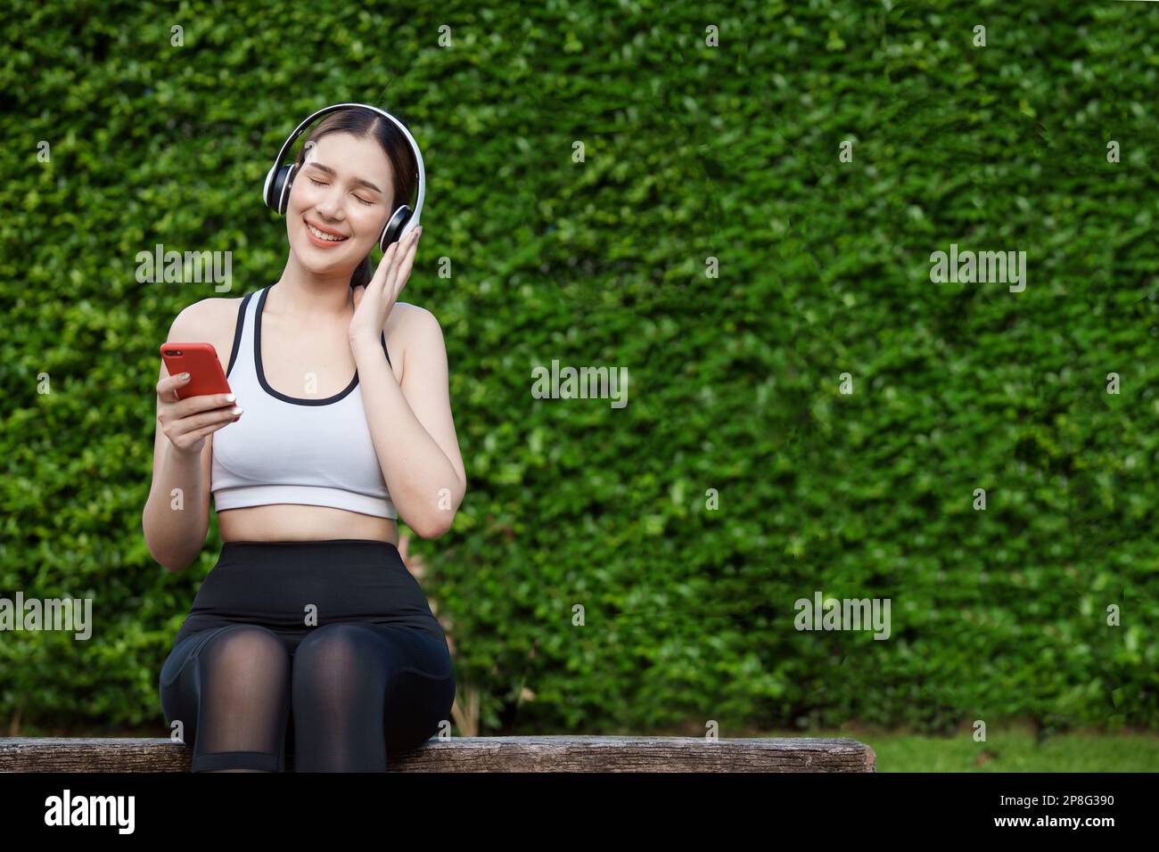 Young woman in sportswear listening music with headphones on  park. She sitting on a bench while resting and using smartphone. Stock Photo