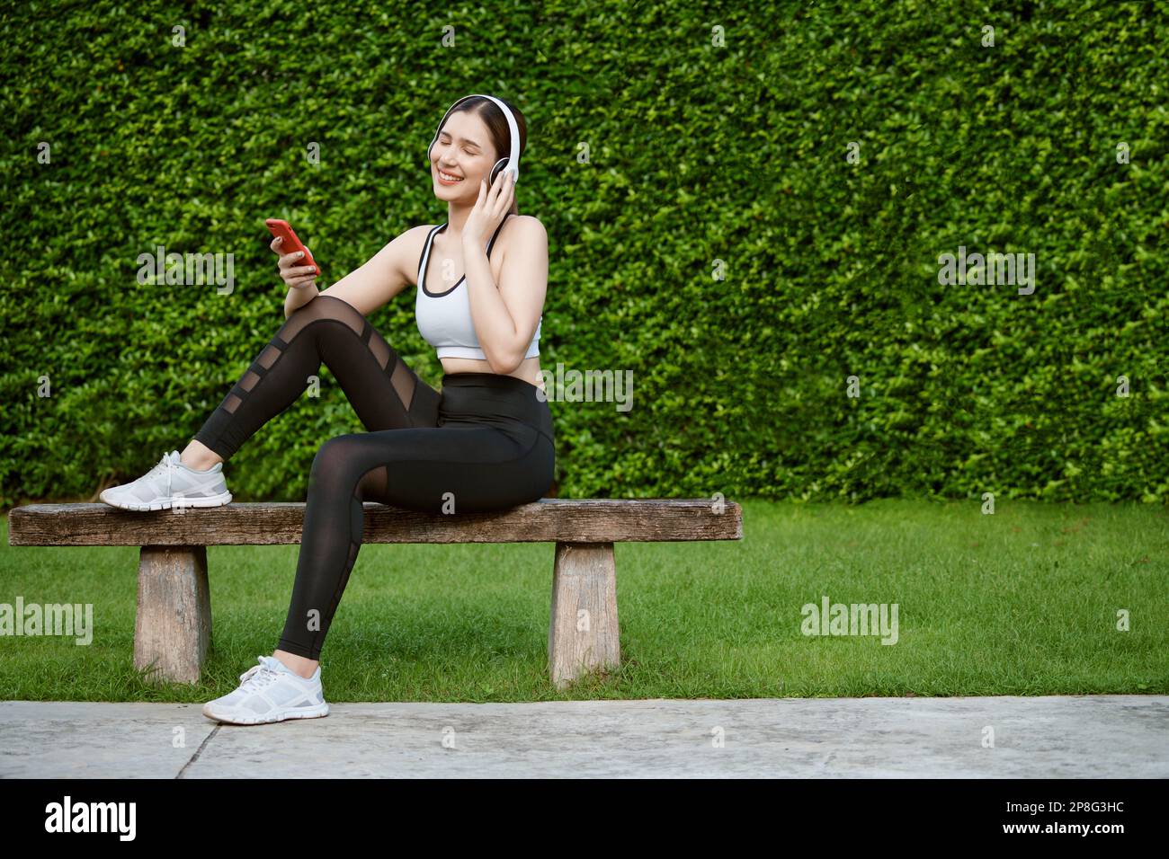 Young woman in sportswear listening music with headphones on  park. She sitting on a bench while resting and using smartphone. Stock Photo