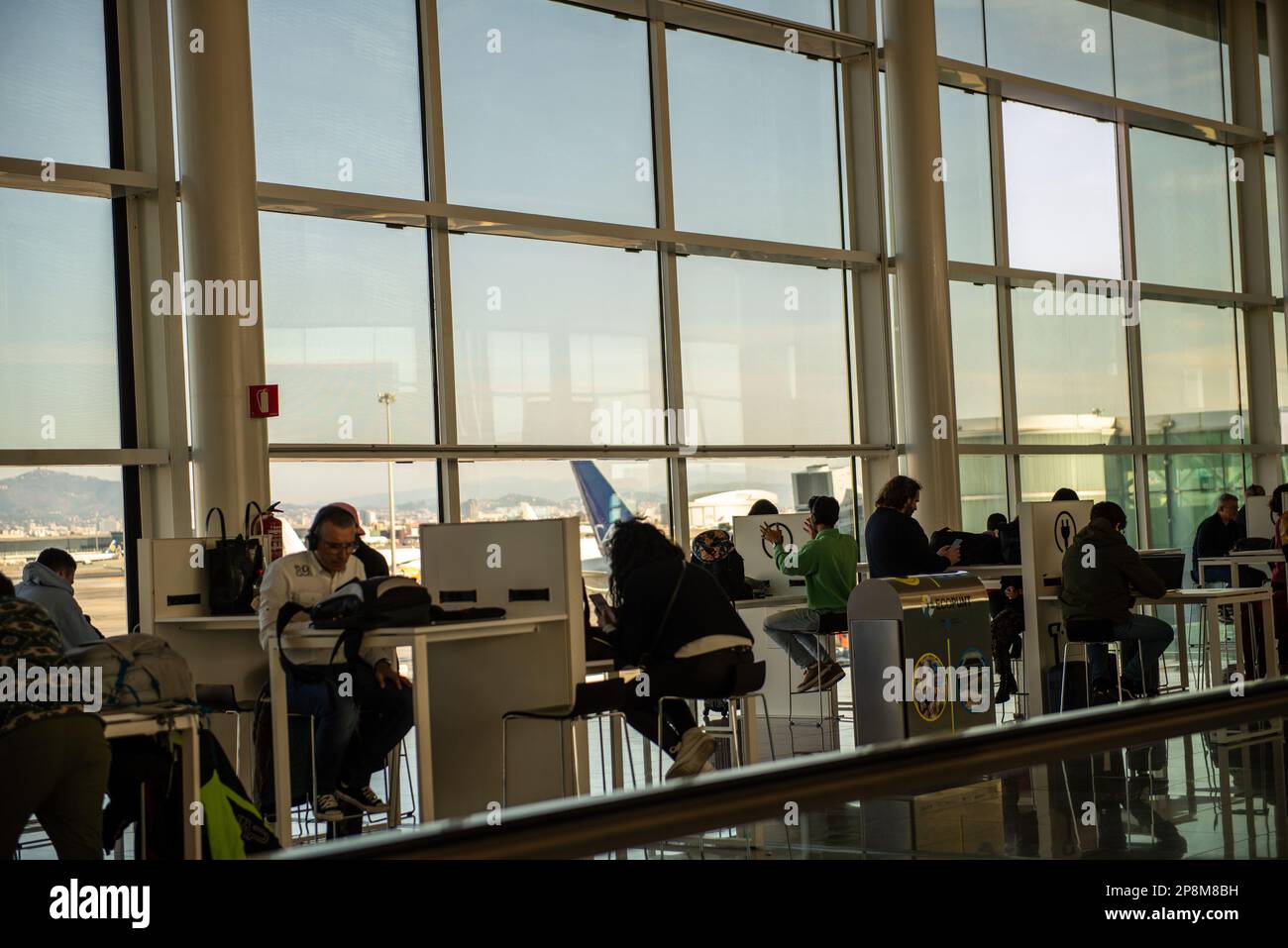 Barcelona,Spain- February 24,2023:People sit at tables by a window charging their smartphones and notebook computers. Stock Photo