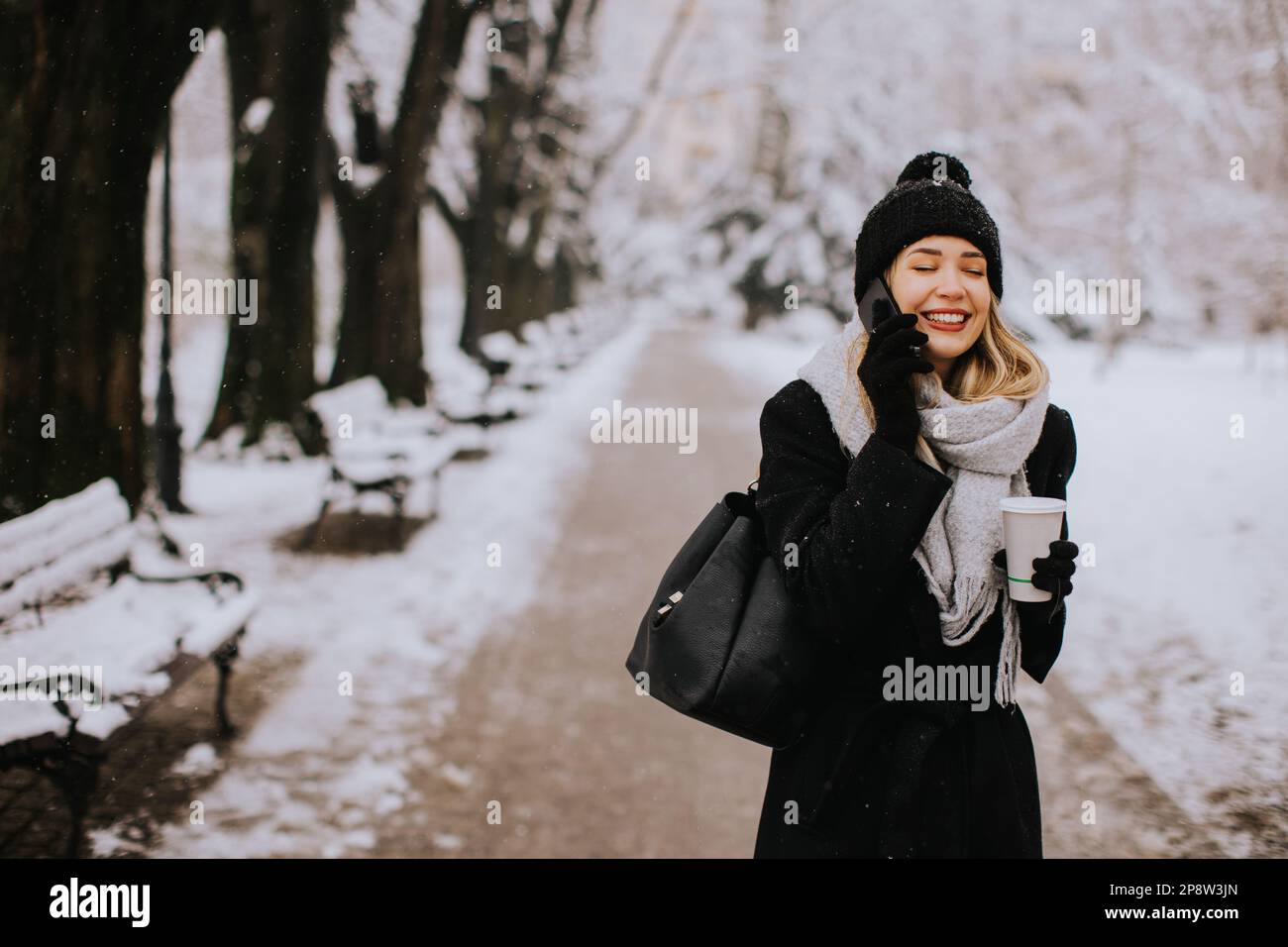 A young woman wearing warm winter clothes and a knit hat smiles happily as she stands in the snow and using mobile phone whil holding cofee cup Stock Photo