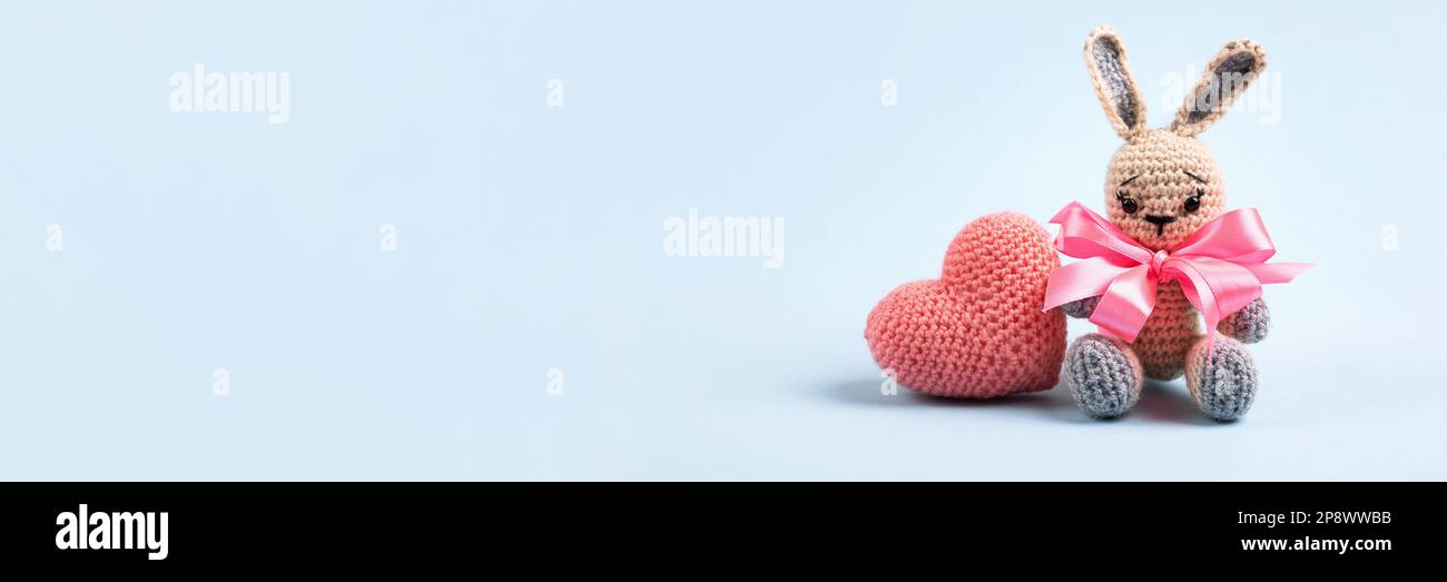 Knitted gray bunny with a crocheted pink heart on a blue background. Banner Stock Photo