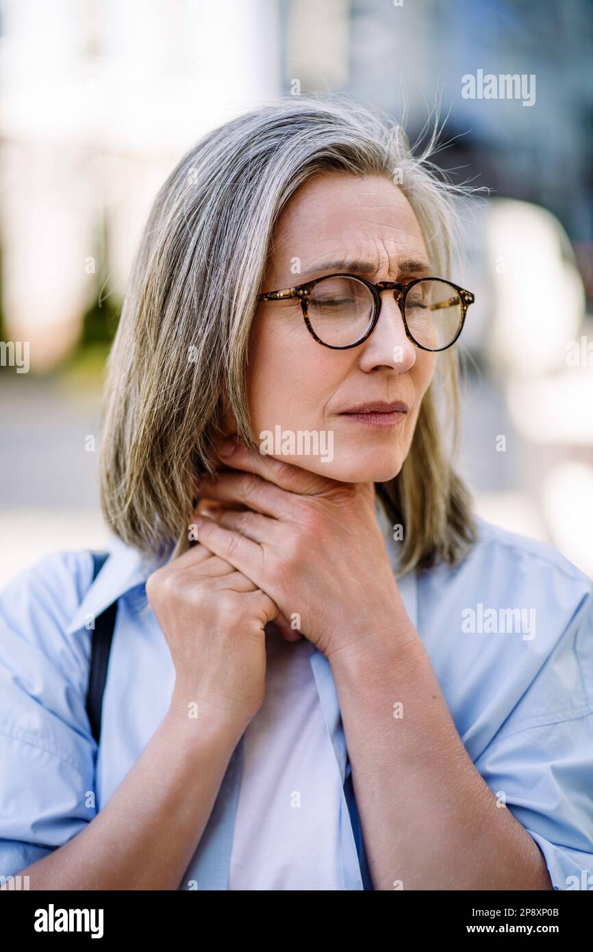 Sad senior woman wearing glasses holding her sore throat outdoors