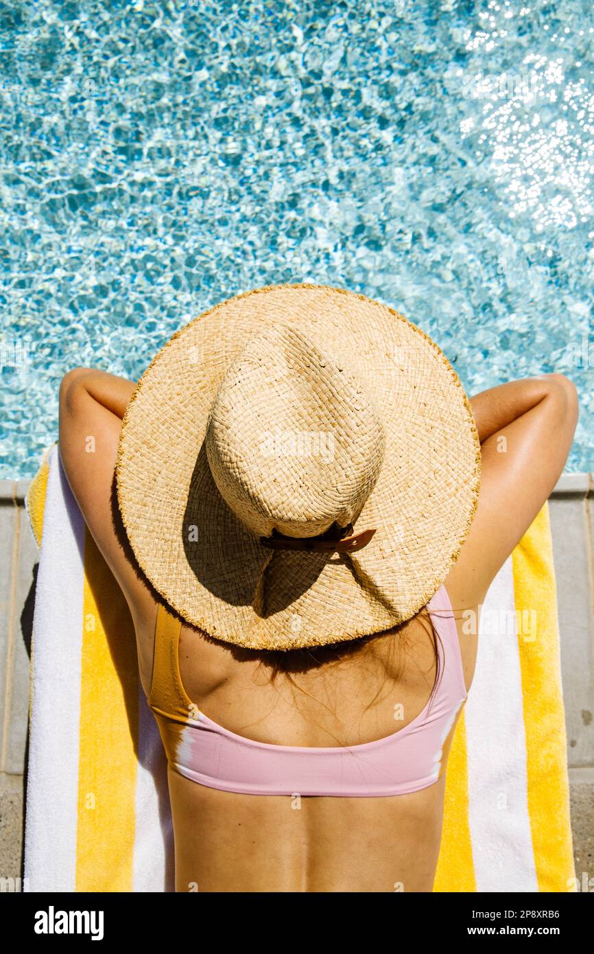 A beautiful woman on vacation lays on a chaise lounge next to a blue pool wearing a sun hat. She is resting her head in her crossed arms gazing out. Stock Photo