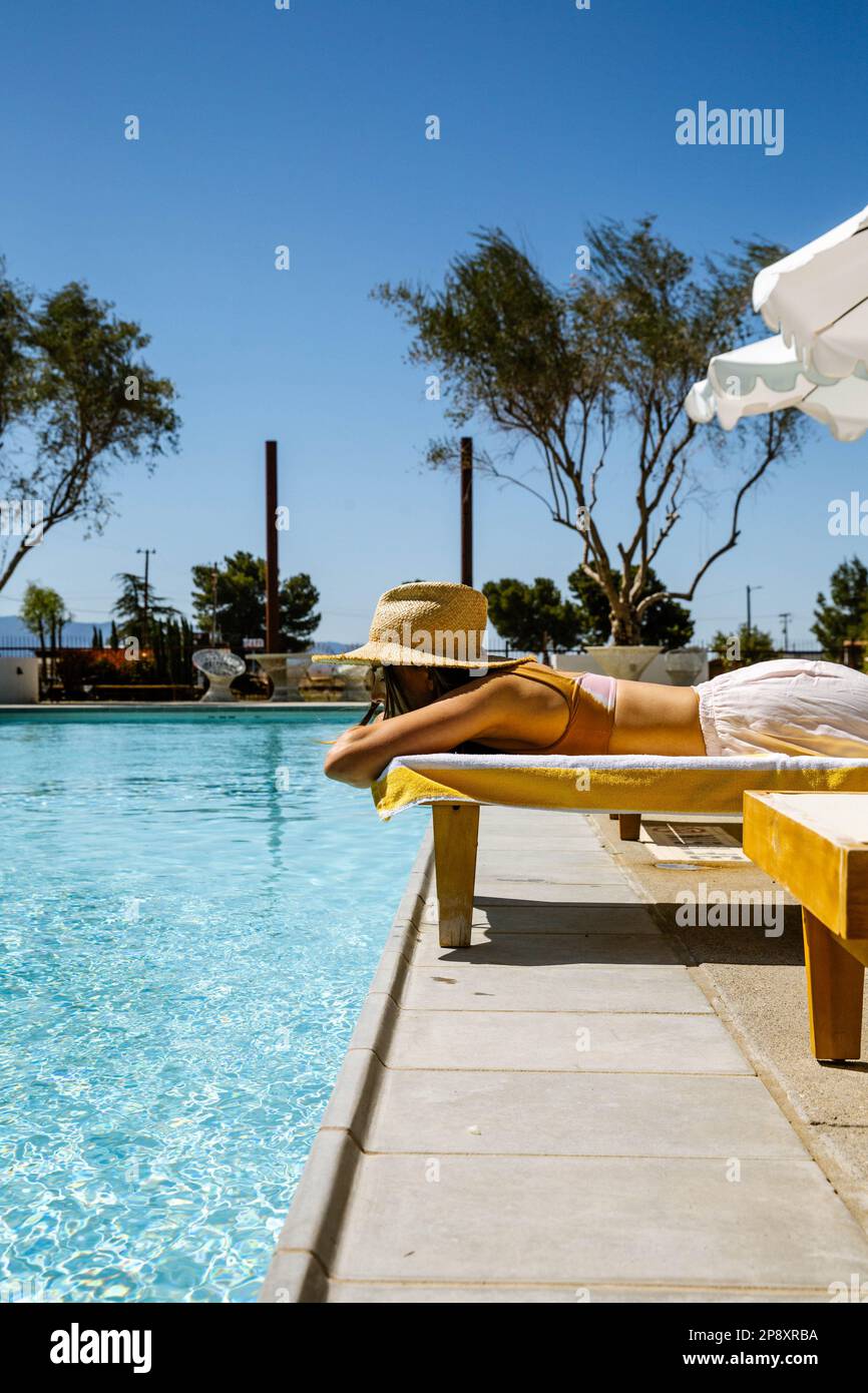 A beautiful woman on vacation lays on a chaise lounge next to a blue pool wearing a sun hat. She is resting her head in her crossed arms gazing out. Stock Photo