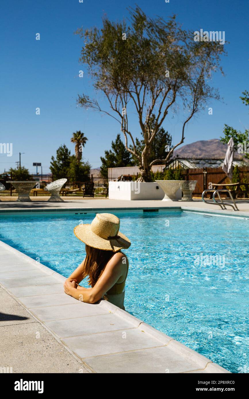 While on vacation, a beautiful woman stands in a blue hotel pool on a sunny day, resting her arms at the edge while looking away. Stock Photo